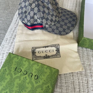 Gucci keps med monogram och läderdetalj - Exklusiv Gucci keps i beige och blått med klassiskt GG-monogram över hela kepsen. Kepsen har justerbar rem i mörkblått skinn och en röd-blå rand på sidan. Kommer med originalbox och dustbag. Perfekt för dig som vill sticka ut med lyxig streetstyle.