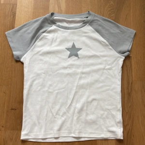 Vit t-shirt med grå stjärna y2k - Y2K Vit t-shirt med grå raglanärmar och en stor grå stjärna tryckt mitt på bröstet. Klassisk rund halsringning och kortärmad modell. Perfekt för en chill och stilren look.