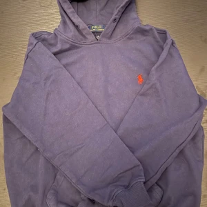 Mörkblå hoodie från Polo Ralph Lauren - Säljer en mörkblå hoodie från Polo Ralph Lauren i klassisk modell med huva och ribbade muddar. Tröjan har det ikoniska röda Polo-logot broderat på bröstet. Perfekt för dig som gillar stilrena och tidlösa plagg med premiumkänsla.