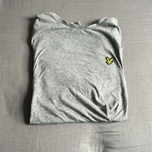 Grå t-shirt från Lyle & Scott - Snygg grå t-shirt från Lyle & Scott med klassisk gul logga på bröstet. T-shirten är i mjuk bomull och har en enkel, stilren design med rund hals och korta ärmar. Perfekt för en chill och avslappnad look. Hör gärna av er vid minsta lilla fråga 