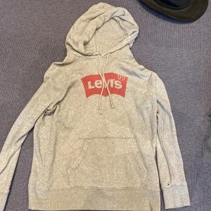 Grå hoodie från Levi's med logga - Grå hoodie från Levi's i mjuk bomullsblandning. Klassisk röd Levi's-logga på bröstet, smått slitage vid ärmmuddarna.