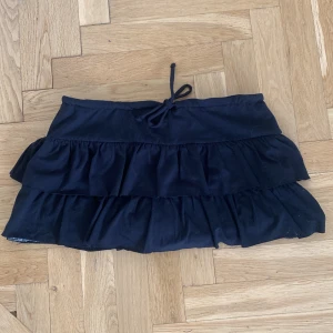 Svart volangkjol med snörning - Supersöt svart kortkjol med dubbla volanger och snörning i midjan. Den är i nyskick och har stretchigt material. Endast använd en gång. Kjolen har även inbyggda shorts.