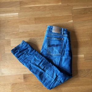Blå jeans från Nudie Jeans - Säljer ett par klassiska blå jeans från Nudie Jeans med snygga slitningar och tydliga sömmar. Jeansen har en normal passform och raka ben, samt Nudie-loggan på bakfickan. Perfekta för dig som gillar en tidlös denimlook.