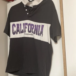 Svart och vit t-shirt från H&M - Cool t-shirt från H&M i svart och vitt med stor lila 'CALIFORNIA' print över bröstet. T-shirten har korta ärmar, vit krage och knappar vid halsen. Tillverkad i 100% bomull för en skön känsla. Perfekt för dig som gillar sportig och avslappnad stil. Säljer för otroligt lågt pris 👍