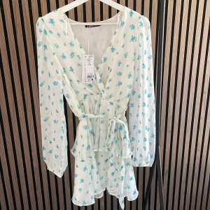 Vit blommig omlottklänning Gina Tricot - Supersöt kort vit klänning från Gina Tricot med blått blommönster, omlottmodell och knytband i midjan. Långa ärmar med lätt puff och volangdetaljer nertill ger en romantisk vibe. (Prislapp på, så helt oanvänd).