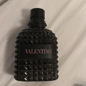 Valentino Uomo Eau de Toilette 100ml - Snygg parfymflaska från Valentino som rymmer 100 ml, flaskan är ca 3/4 full. Flaskan är svart med nitar och har en lyxig, edgy design i glas. Märket står i rosa bokstäver på svart etikett. Perfekt för dig som gillar exklusiva dofter och coola detaljer.