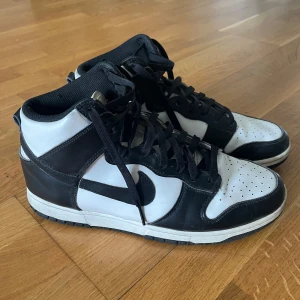 Nike Dunk High storlek 43 - Säljer ett par klassiska Nike Dunk High i svart och vitt skinn. Skorna har svart swoosh, svarta snören och vit mellansula. Perfekta för dig som gillar streetwear och vill ha en ikonisk sneaker med hög siluett.