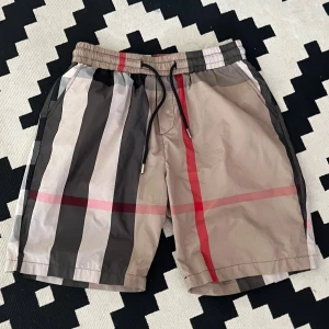 Burberry-rutiga shorts med snörning - Snygga shorts från Burberry med klassiskt rutigt mönster i beige, svart, vitt och rött. De har elastisk midja med snörning och är tillverkade i ett lätt syntetmaterial, perfekta för sommaren. Stilen är loose och de har en modern vibe som sticker ut.