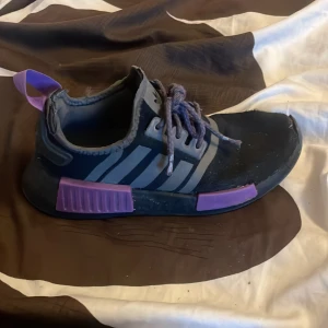 Adidas NMD sneakers svart/lila - Säljer ett par Adidas NMD sneakers i svart med gråa ränder och lila detaljer på sula och hälflik. Skorna har snörning och en sportig, modern design med platt sula. Ovandelen är i syntetmaterial och mesh för en skön känsla.