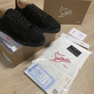 Christian Louboutin svarta sneakers med nitar - Svarta sneakers från Christian Louboutin i mocka med svarta nitar på tån och klassisk röd sula. Skorna har snörning och en stilren, rund tå. Perfekta för dig som vill sticka ut med exklusiva detaljer och edgy vibe.