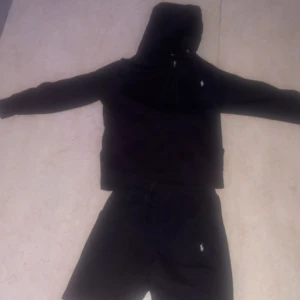 Svart hoodie från Polo Ralph Lauren - Svart hoodie från Polo Ralph Lauren med klassisk vit broderad logga på bröstet. Jackan har huva med snörning, dragkedja framtill och känguruficka. Tillverkad i mjuk bomull, perfekt för chill dagar eller när du vill ha en clean streetstyle.