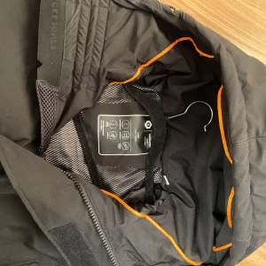 Svart dunjacka Jack & Jones L - Svart dunjacka från Jack & Jones med huva och orange detalj på insidan. Jackan har dragkedjor på fickorna, bröstficka och storm cuffs. Vindtät och vattenavvisande med teknisk vaddering, perfekt för kalla dagar.