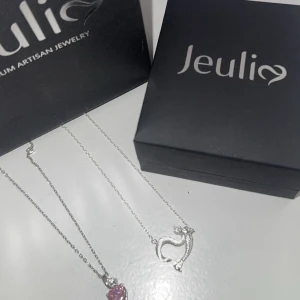 Jeulia halsband med katt och hjärta - Två fina halsband från Jeulia. Ena har ett silverfärgat hänge i form av en katt och det andra ett glittrigt rosa hjärta. Båda har tunna kedjor i silverfärg och levereras i originalask. Perfekt för dig som älskar söta detaljer och djurmotiv.