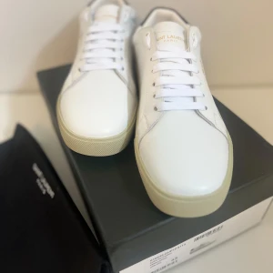 Vita Saint Laurent sneakers i skinn - Snygga vita sneakers från Saint Laurent i slätt skinn med guldtext på sidan. Klassisk låg modell med vit sula och vita skosnören. Diskret svart detalj på hälen och rund tå. Perfekta för dig som gillar stilrena och exklusiva sneakers.