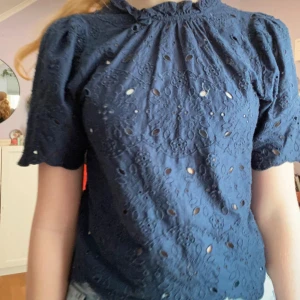 Mörkblå blus med broderade blommor - Supersöt mörkblå blus med broderade blommor och hålmönster. Puffiga korta ärmar och rundad halsringning med liten volangkant. Blusen har vågig nederkant och är perfekt till jeans eller kjol. Materialet känns lätt och luftigt.