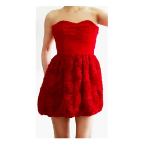 Röd axelbandslös kortklänning - Supersöt röd axelbandslös kortklänning med hjärtformad överdel och voluminös kjol med rosdetaljer. Perfekt för dig som vill sticka ut och känna dig extra fin. Kjolen har en fluffig struktur och överdelen är figurnära.