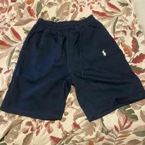 Mörkblå shorts från Polo Ralph Lauren - Snygga mörkblå shorts från Polo Ralph Lauren med klassisk vit logga broderad på benet. De har elastisk midja med snörning för skön passform och är tillverkade i mjuk bomull. Perfekta för chill dagar eller när du vill ha en clean och stilren look.