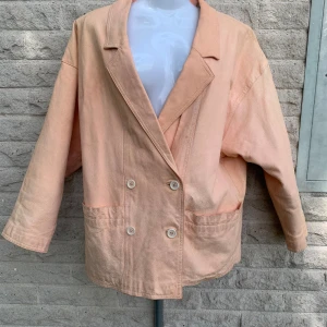 Ljusrosa vintage skinnkavaj i svinnappa - Oversized kavaj i ljusrosa svinnappa med dubbelknäppning och stora fickor framtill. Jackan har klassisk krage, raka ärmar och är fodrad med bomull. Perfekt för dig som gillar en chill och trendig look med en twist av vintage.