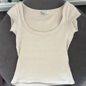 Beige ribbad topp från Zara - Superenkel och snygg beige topp från Zara i ribbad bomull. Modellen har korta ärmar och en bred, rundad urringning som ger en clean look. Perfekt att matcha med jeans eller kjol för en chill stil.