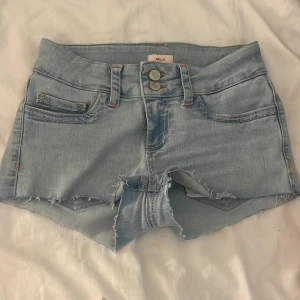 Ljusblå jeansshorts från Nelly storlek 34 - Snygga ljusblå jeansshorts från Nelly med råa kanter och dubbla knappar framtill. Shortsen har lite mönster på backfickan.Dom är helt oanvända. Storlek 34