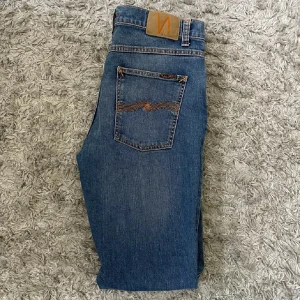 Blå jeans från Nudie Jeans - Snygga blå jeans från Nudie Jeans i klassisk modell med orangea sömmar och ikonisk bakficka. Jeansen har normal passform och raka ben, tillverkade i slitstarkt bomullsjeans. Perfekta för dig som gillar stilrena och tidlösa jeans.