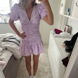 Ljuslila kort klänning med volang - Superfin ljuslila kort klänning med broderade blommor, puffiga kortärmar och v-ringning. Klänningen har rynkade detaljer över hela livet och en volangkant nertill. Perfekt för sommarens alla tillfällen!