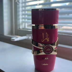 Yara parfym från Lattafa - Snygg parfymflaska i starkt rosa med gulddetaljer och dekorativa band runt flaskan. Märket Lattafa och namnet Yara syns tydligt i guldtext. Flaskan har en modern, lyxig design som sticker ut på hyllan. Har använt några gånger men är helt full.