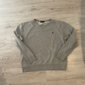 Grå sweatshirt från Polo Ralph Lauren - Klassisk grå sweatshirt från Polo Ralph Lauren med broderad logga på bröstet. Tröjan har rund hals, långa ärmar och ribbade muddar. Perfekt för en chill och stilren look. Tillverkad i mjuk bomull för extra komfort.