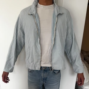 Ljusblå jacka med dragkedja - Säljer en stilren ljusblå vindjacka med diskret randigt mönster. Jackan har hög krage, hel dragkedja framtill och långa ärmar. Perfekt för dig som gillar en clean och enkel look. Passar till jeans eller chinos för en avslappnad vibe.