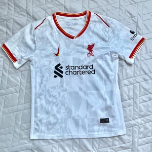Liverpool Salah 11 bortatröja Nike - Liverpool FC bortatröja med M. Salah 11 på ryggen. Vit med subtilt mönster, röda och gula detaljer på krage och ärmslut. Nike-logga och klubbmärke framtill, Standard Chartered sponsortryck. Lätt och ventilerande material, perfekt för match eller träning. Har använt ca 5 ggr och passar folk från ca 155-160.