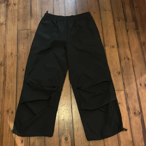 Svarta parachute pants - Svarta täckbyxor med elastisk midja och dragsko vid benslut. Byxorna har en loose passform och är perfekta för kalla dagar. Tillverkade i ett syntetmaterial som står emot väta och vind. Nytt skick.