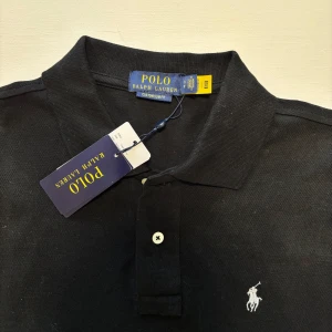 Ralph Lauren Piké - Klassisk svart pikétröja från Polo Ralph Lauren med vit broderad logga på bröstet. Tröjan har korta ärmar, krage och två knappar framtill. Slim fit-modell i mjuk bomull som ger en clean och stilren look. Perfekt för sommar dagar! Skickas inom 24 timmar! Storleken är S, kan även passa någon med M. 