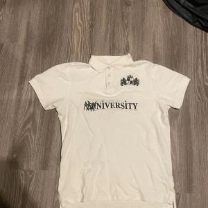 Aint Bout Nun limited university polo vit - ABN AINT BIUT NUN Vit pikétröja med svart tryck där det står 'University' och ett emblem på bröstet. Klassisk krage med knappar och korta ärmar. Perfekt för en clean och stilren look. Materialet känns som bomull och tröjan har en normal passform.