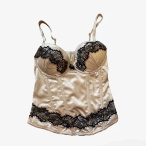 Champagnefärgad korsett med spets - Supersnygg korsett i champagnefärgad satin med svarta spetsdetaljer över bysten och längs nederkanten. Axelbanden är justerbara och modellen har markerade sömmar för en formad look. Perfekt för dig som vill sticka ut lite extra.