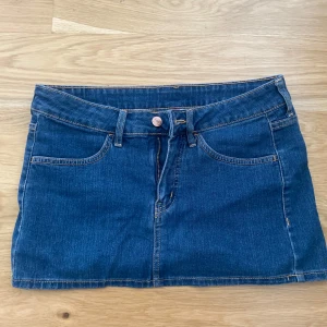 Blå jeanskjol från H&M Divided - Snygg blå jeanskjol från H&M Divided med klassisk femficksdesign, bälteshällor och dragkedja framtill. Kjolen är kort och har en rak passform som passar perfekt till sneakers eller boots. Perfekt för dig som gillar en enkel men trendig stil.