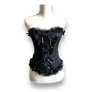 Svart korsett med spets och snörning - Supersnygg svart korsett med dekorativ spetskant upptill och nertill samt detaljerad snörning framtill. Sidenliknande tyg med broderade band och ögonhål för snörningen. Perfekt för dig som vill ha en edgy och cool look med romantiska vibbar.