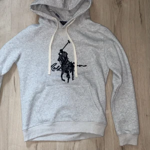 Grå hoodie från Polo Ralph Lauren - Snygg grå hoodie från Polo Ralph Lauren med klassisk svart polospelare på bröstet. Tröjan har huva med vita snören och en stor magficka. Perfekt för dig som gillar streetstyle och vill ha en chill look.