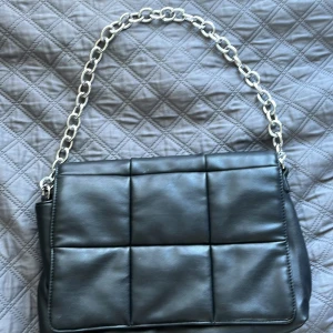 Svart quiltad axelväska med kedja - Snygg svart axelväska med quiltad design och en rejäl silverfärgad kedja som axelrem. Väskan har en stilren, rektangulär form och är gjord i skinnimitation. Perfekt för dig som vill ha en trendig och praktisk väska med coola detaljer.