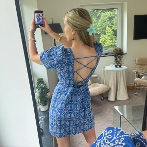 Blå kortklänning med snörning i ryggen - Superfin blå kortklänning med vitt blommigt mönster, v-ringning och puffärmar. Ryggen är öppen med snygg snörningsdetalj. Perfekt för sommarens alla tillfällen och ger en fräsch vibe. Materialet känns lätt och luftigt. Använd 1 gång 