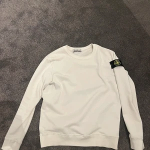 Vit sweatshirt från Stone Island - Vit långärmad sweatshirt från Stone Island med klassisk rund hals och ikonisk svart logotyp-patch på vänster ärm. Tröjan har en clean look och är tillverkad i mjuk bomull, perfekt för en stilren och avslappnad vibe.