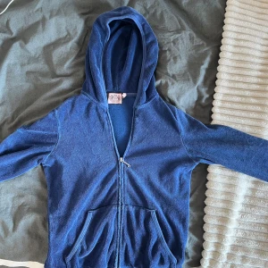 Blå velour hoodie från Juicy Couture - Blå velour hoodie från Juicy Couture med dragkedja framtill, huva och kängurufickor. Baksidan har glittrande Juicy-tryck och dekorativa detaljer. Har dock 2 små hål i fickorna. Läs min bio!