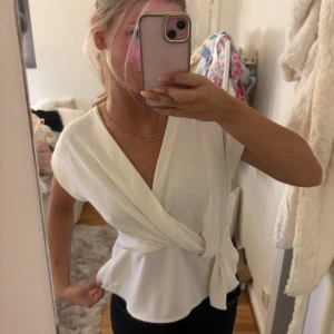 Vit omlottopp med knyt i midjan - Säljer en stilren vit omlottopp med v-ringning och knyt i midjan. Toppen har korta ärmar och ett flowy fall som ger en snygg siluett. Perfekt att matcha med jeans eller kjol för en fräsch look.