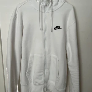 Vit Nike hoodie med dragkedja/ byte  - Snygg vit hoodie från Nike med svart broderad logga på bröstet. Tröjan har hel dragkedja, huva med snörning och två stora fickor framtill. Perfekt för en chill och sportig look. Tillverkad i mjuk bomullsmix för extra komfort.