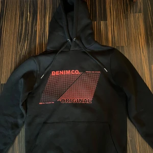 Svart hoodie från SMOG med tryck - Svart hoodie från SMOG i regular fit med rött grafiskt tryck och texten 'DENIM CO. ORIGINAL' på bröstet. 140 kr + frakt, eller 100 kr vid upphämtning på Hulta. Den är använd bara TVÅ gånger! 