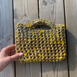 Guldig handväska med flätad look - Unik handväska i två olika nyanser av guld samt gult med flätad design och inbyggt handtag. Väskan är virkad och har en cool, chunky känsla. Perfekt för dig som vill sticka ut med accessoarer i metallic och färg. Väskan är 22cm bred och 15cm hög