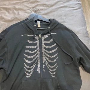 Svart hoodie med skelettmönster från Independent Trading Company - Cool svart hoodie med dragkedja från Independent Trading Company. Framsidan har ett glittrigt skelettmönster i silver. Tröjan har huva med snören och fickor framtill. Perfekt för dig som gillar unika detaljer!