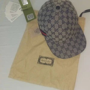 Gucci keps med monogrammönster - Snygg keps från Gucci i blått och beige med klassiskt GG-monogram över hela kepsen. Justerbar rem baktill i mörkblått skinn. Kepsen är tillverkad i bomull och har märkets logga invävd i tyget. Tjena säljer min gucci keps efter som att den inte kommer till användning längre. Köpts i Danmark 2023 tyvärr har jag inte kvittot och boxen kvar efter som att jag hade flyttade 1år sedan och tappade bort både boxen och kvittot