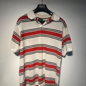 Randig pikétröja i rött och vitt - Snygg pikétröja med breda röda, svarta och vita ränder. Klassisk krage och korta ärmar. Tröjan är i bomull och har en skön passform som funkar året runt.