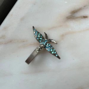 Silverfärgad ring med blå stenar - Unik ring i silverfärgad metall med en stor fågeldekoration. Fågeln har vingar täckta av små blå glittrande stenar som ger en cool och iögonfallande look. Perfekt accessoar för dig som vill sticka ut med något annorlunda och snyggt.