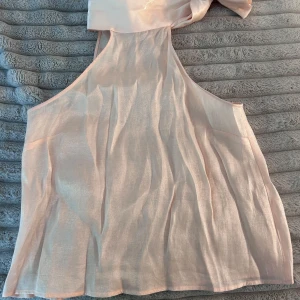 Ljusrosa omlottopp från MNG - Supersöt ljusrosa omlottopp från MNG i glansigt och tunt material. Toppen har en bred knytning vid halsen som ger en snygg detalj och är ärmlös med luftig passform. Perfekt för dig som gillar en feminin och trendig look.